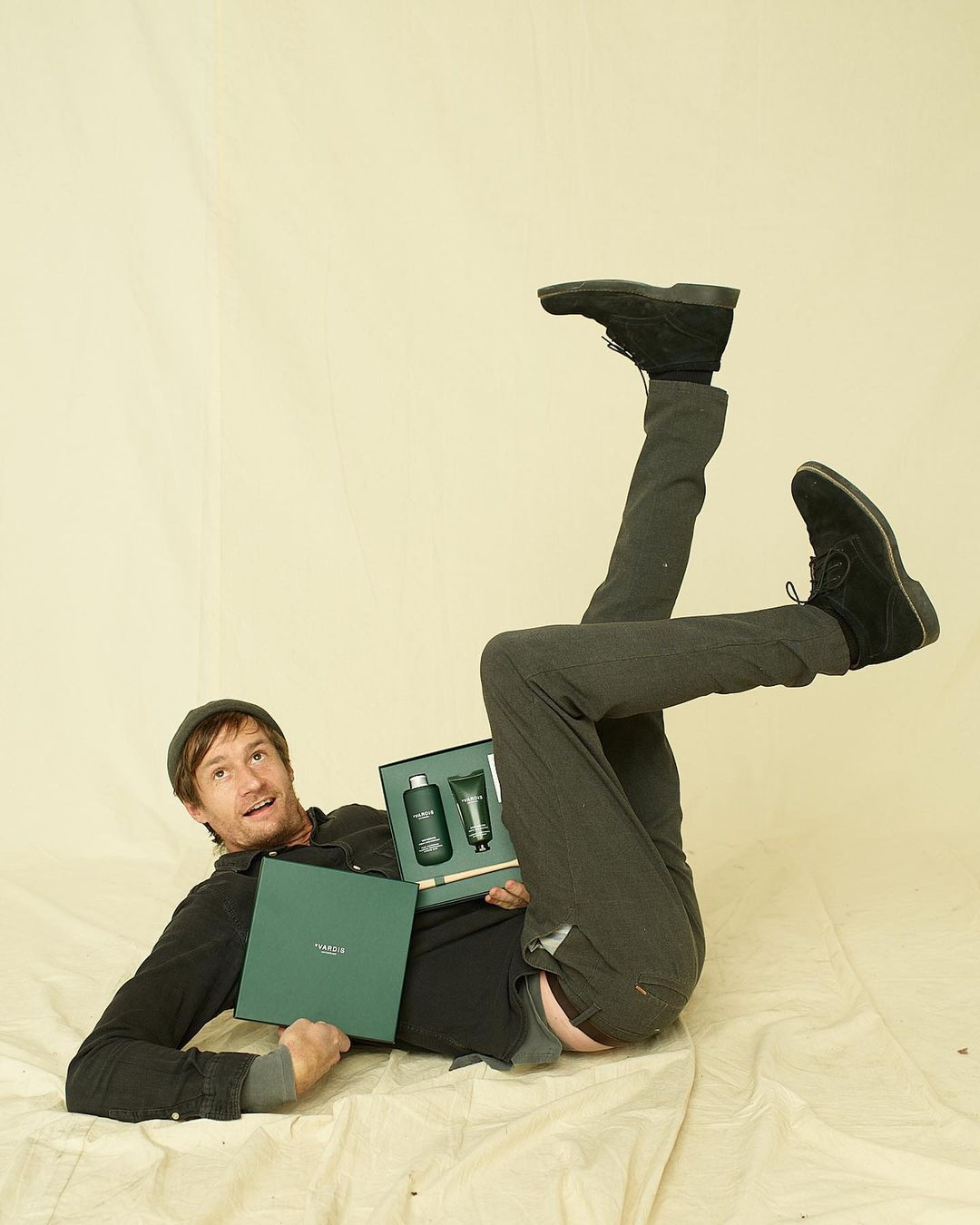 Man lying on his back holding a skincare gift set, wearing black shoes, grey pants and shirt, shot in a photo studio on a white backdrop.