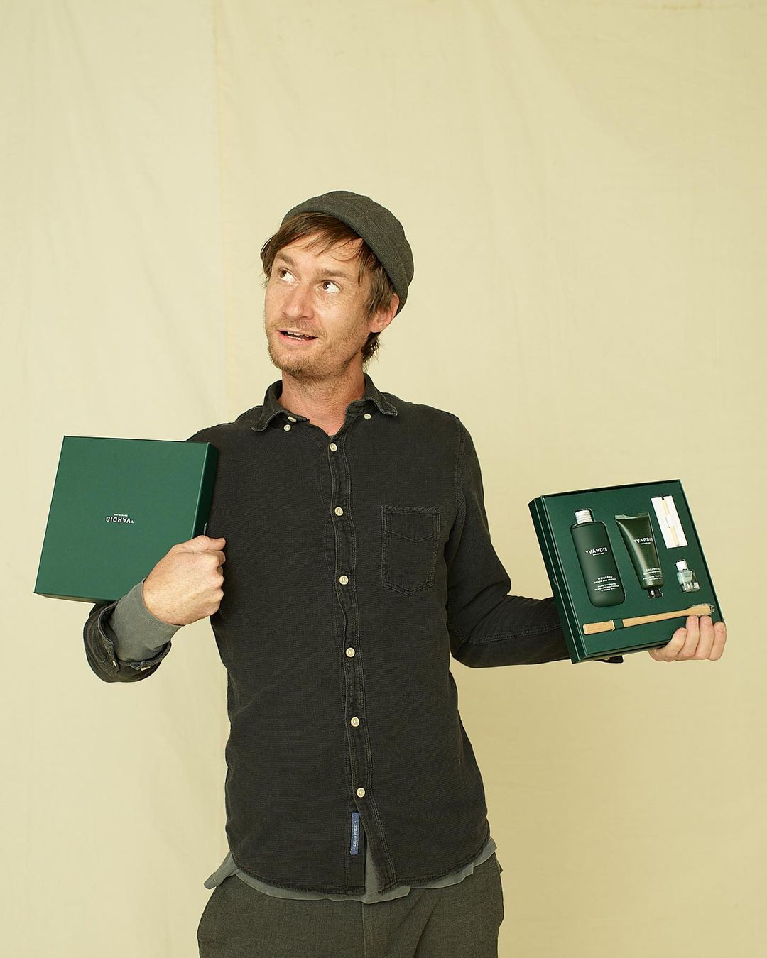 Adult male with brown hair and gray cap holding two green boxed sets of men's skincare products against a tan background.