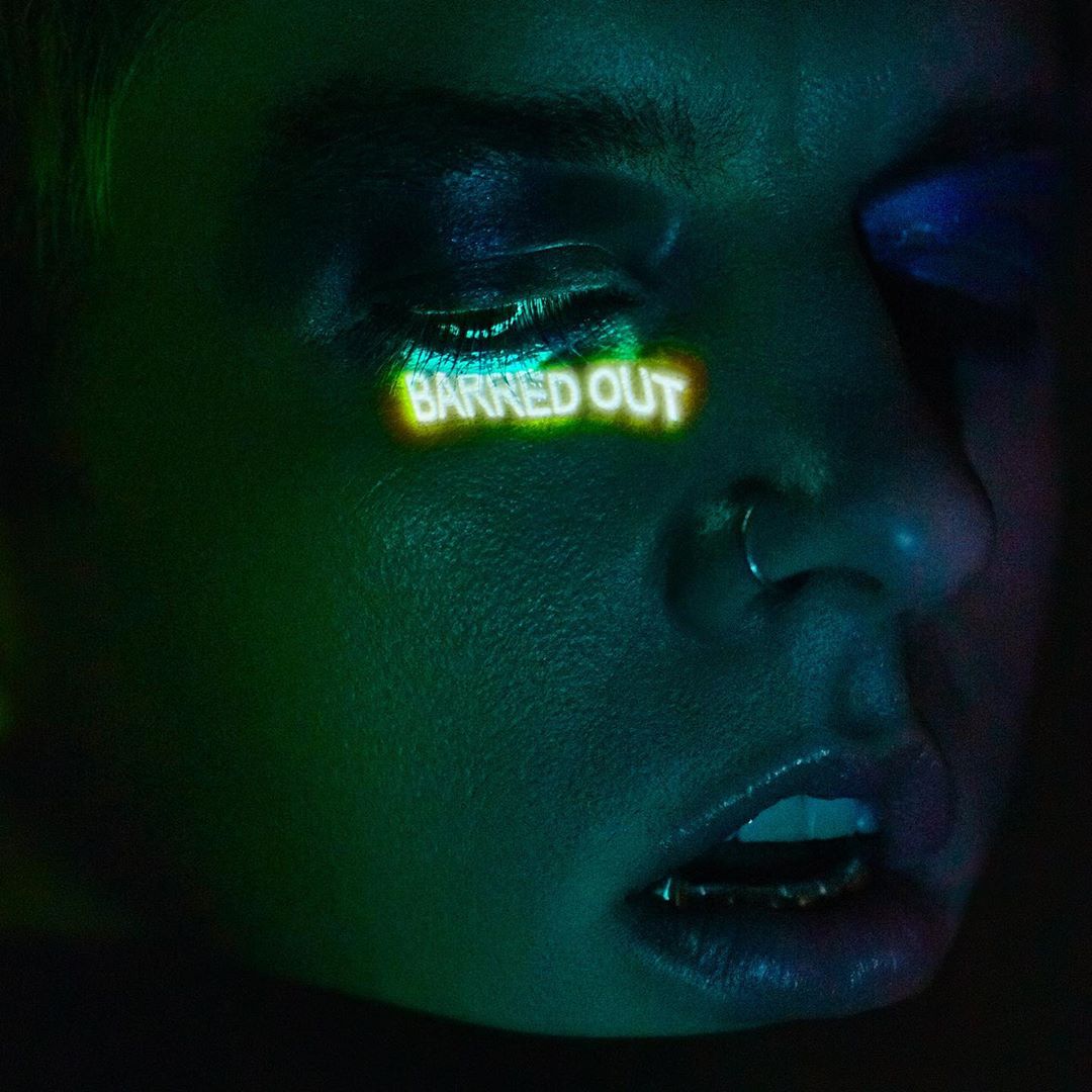 Close-up portrait of an adult female with dramatic blue and purple eyeshadow, nose ring, and a reflective golden grill on her lower teeth.