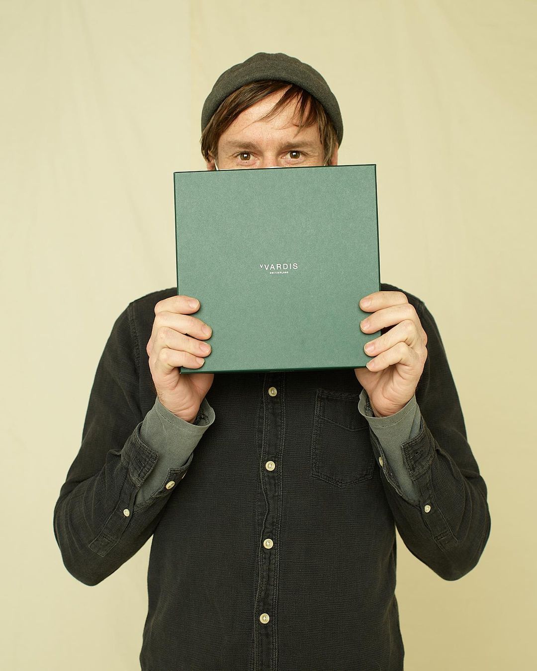An adult man in a gray beanie holding a square green box with the text 'VARDIA' printed on it in front of his face in a neutral indoor setting.