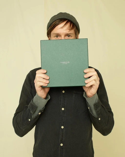 An adult man in a gray beanie holding a square green box with the text 'VARDIA' printed on it in front of his face in a neutral indoor setting.