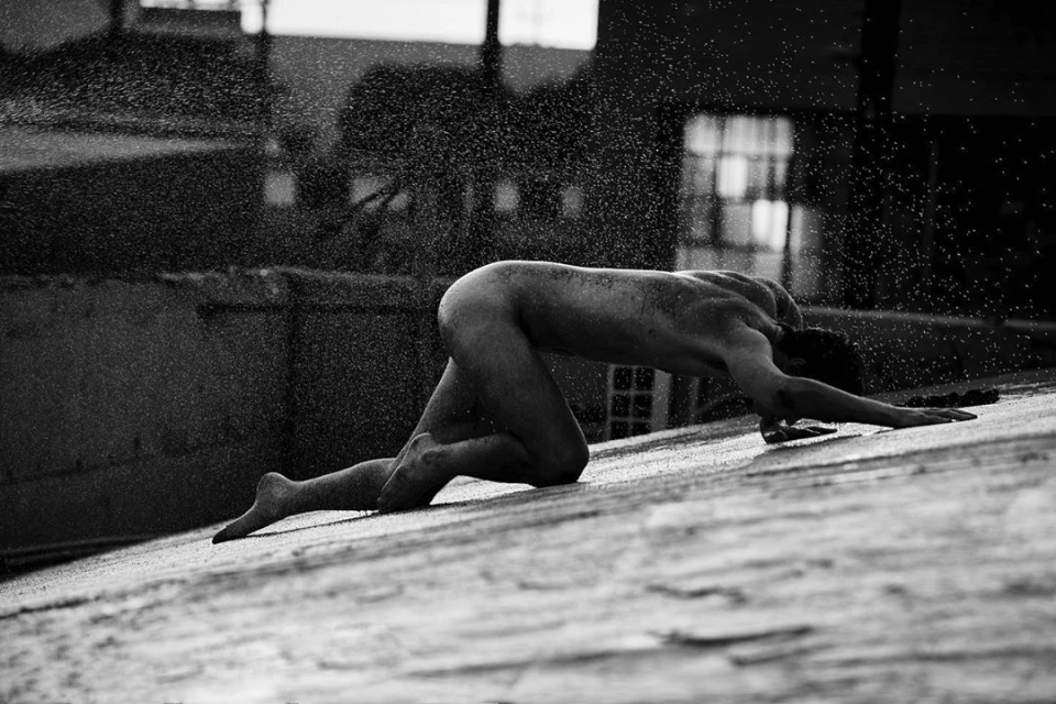 Nude adult male kneels on a flat rooftop surface under water droplets against an out-of-focus cityscape in black and white.