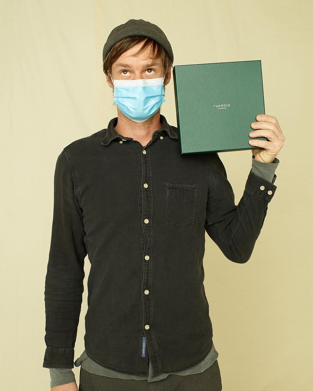 Adult male wearing a face mask and beanie presents a dark green gift box in a studio setting. Casual fashion with soft lighting.