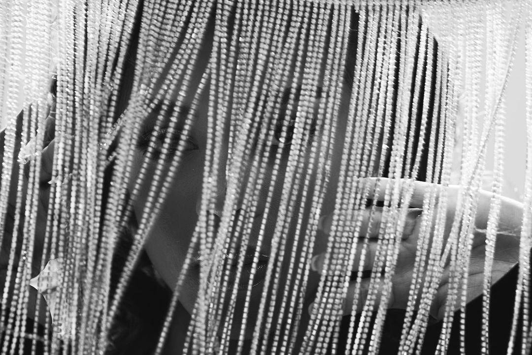 Monochrome portrait of an adult with fair skin partially obscured by a beaded curtain, creating a textured, abstract visual effect.