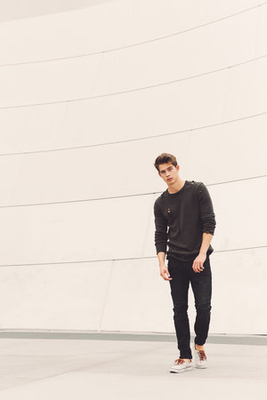 A young man stands against a curving white wall, wearing a charcoal-colored sweater, black jeans, and off-white sneakers, looking at the camera.