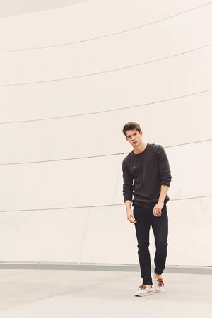 A young man stands against a curving white wall, wearing a charcoal-colored sweater, black jeans, and off-white sneakers, looking at the camera.