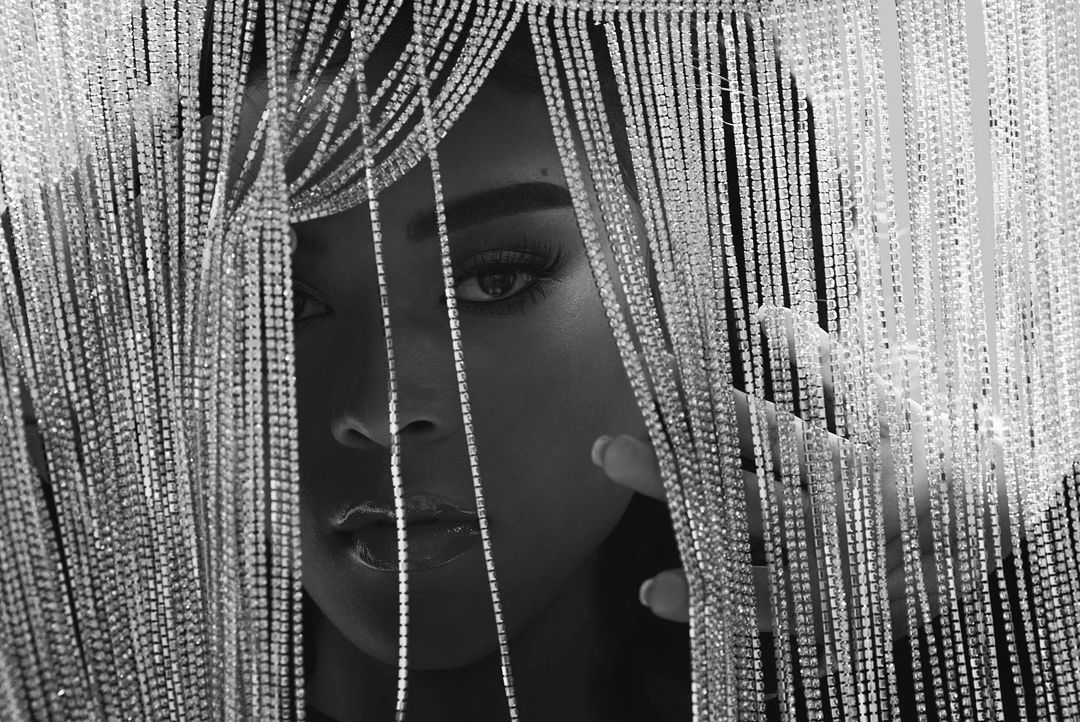 Monochrome portrait of a Black woman with shiny beaded fringe framing her face, partially covering one eye and cheekbone.