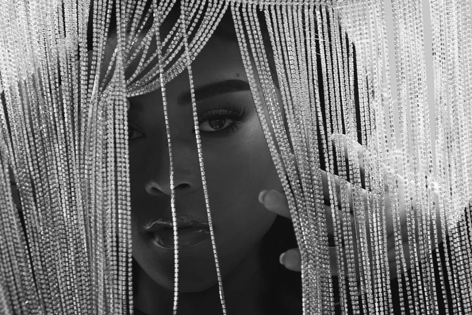Monochrome portrait of a Black woman with shiny beaded fringe framing her face, partially covering one eye and cheekbone.