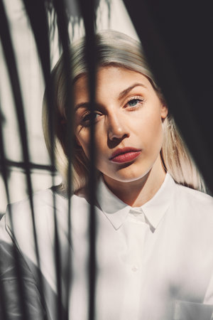 Close-up portrait of a blonde woman wearing a white collared shirt, partially silhouetted by dark vertical shadows.