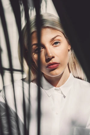 Close-up portrait of a blonde woman wearing a white collared shirt, partially silhouetted by dark vertical shadows.