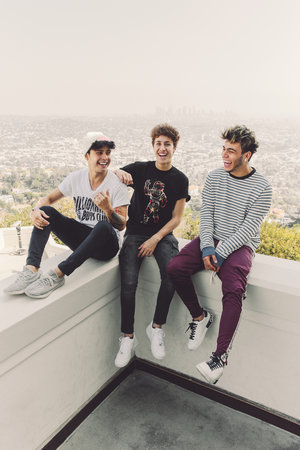 Three young adult men sit on a ledge, smiling and laughing, with a city skyline and hazy sky visible in the distance during daytime.