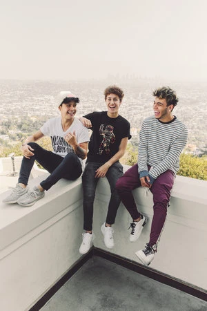Three young adult men sit on a ledge, smiling and laughing, with a city skyline and hazy sky visible in the distance during daytime.