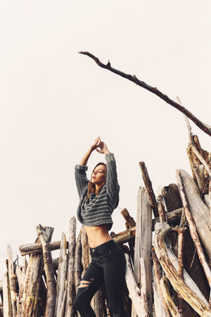 A young woman in ripped jeans and a cropped sweater stands in front of weathered driftwood on a beach with her arms overhead.