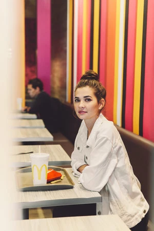 Young woman sitting at a fast-food restaurant table with a drink and food tray. Another patron sits in the background.