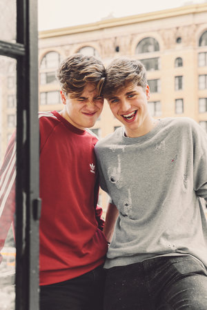 Two young adult men smiling together in an urban environment. The man on the left is wearing a red Adidas shirt, and the other wears grey.