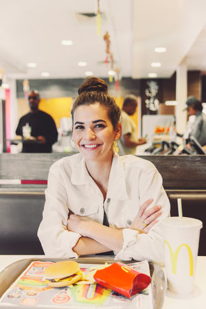 Smiling brunette woman in a white jacket sits at a McDonald's table with a burger, fries, and drink in a brightly lit restaurant.