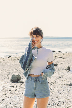 Smiling woman in denim jacket, top, and shorts stands on a rocky beach on a sunny day, looking at the camera.
