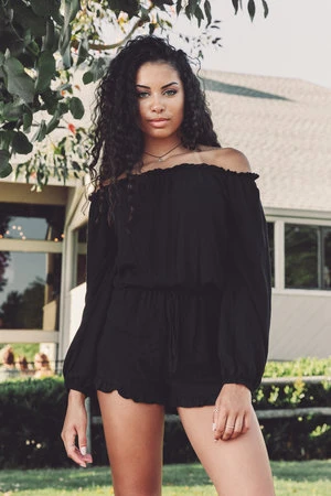 Attractive young woman in a black romper posing outdoors in front of a house with trees, showcasing fashion and style.