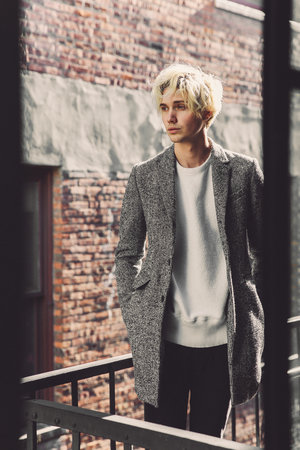 Stylish adult male stands on a balcony with his hands in his pockets, wearing a white sweater, a gray tweed coat, and black pants.