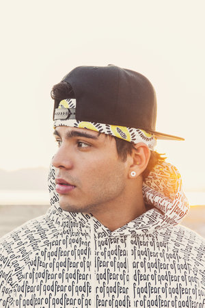Portrait of a young adult male with a black cap, patterned bandana, and hoodie. He looks to the left against a neutral outdoor background.