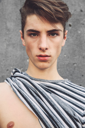 Young adult male in a striped draped top poses against a grey concrete wall, captured in a medium close-up shot with natural lighting.