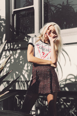 A young woman poses outdoors in a stylish tweed skirt and Guess logo t-shirt, bathed in sunlight and surrounded by greenery.