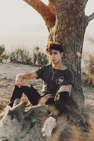 Young man seated at the base of a large tree trunk wearing a dark t-shirt and distressed black jeans.