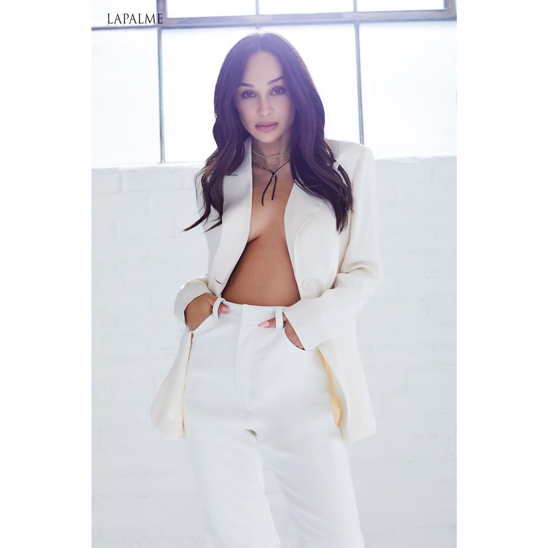 A brunette woman in a white suit poses in front of a window with bright lighting in a fashion-style photo with LaPalme branding.