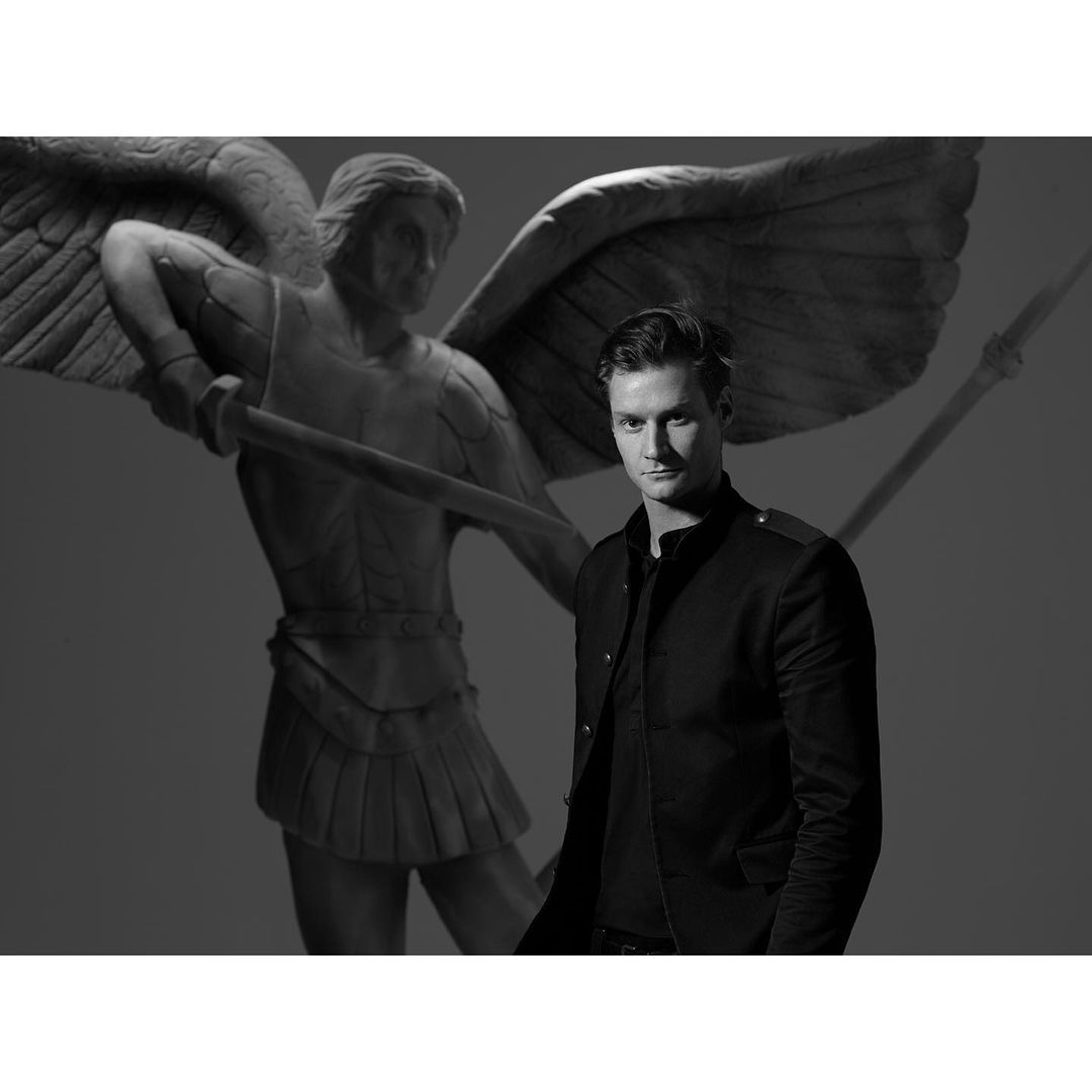 Monochrome portrait of a dark-haired adult male in a black jacket, posing in front of a statue of an angel with wings and a spear.