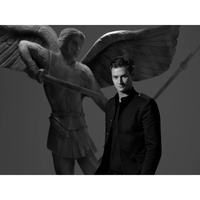 Monochrome portrait of a dark-haired adult male in a black jacket, posing in front of a statue of an angel with wings and a spear.