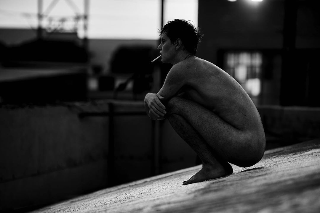 Monochrome image of a shirtless adult male squatting outdoors and smoking a cigarette, looking off into the distance, with an urban background.