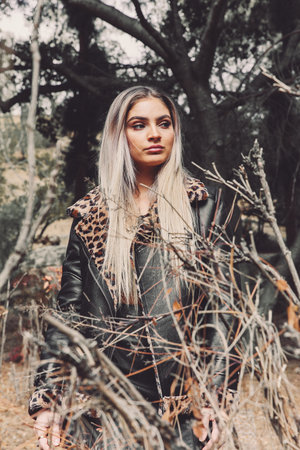Woman in leather jacket with faux fur collar poses outdoors amid bare branches, looking off-camera with neutral facial expression.
