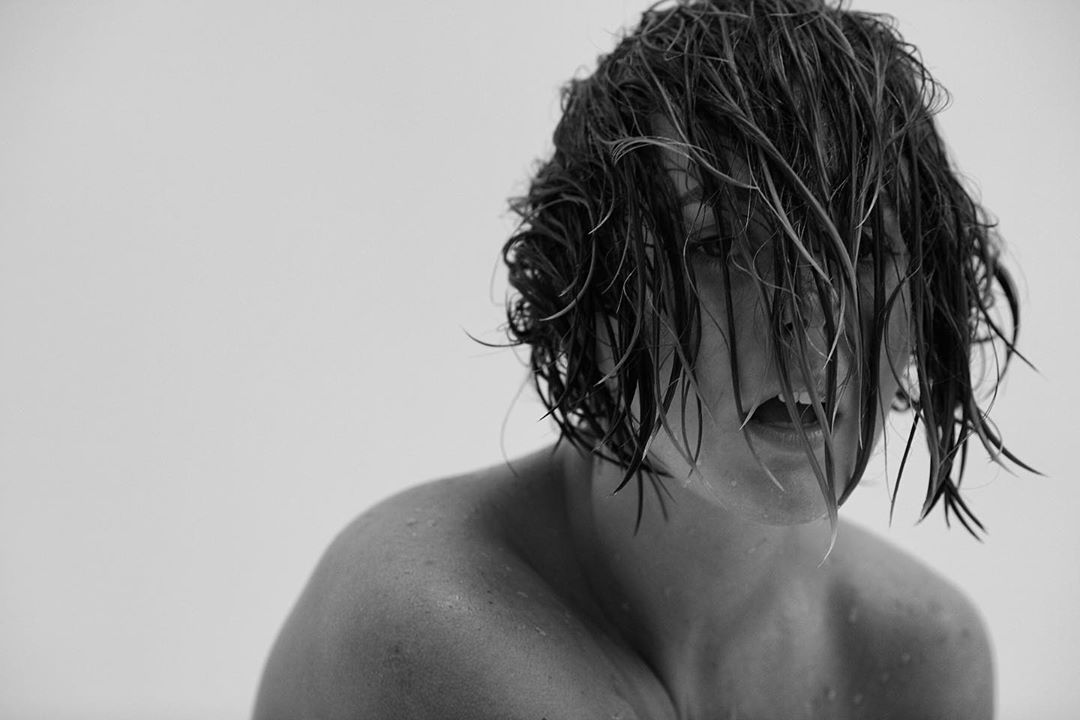 Monochromatic portrait of a person with wet hair partially obscuring their face, shoulders and upper chest visible against a stark white backdrop.