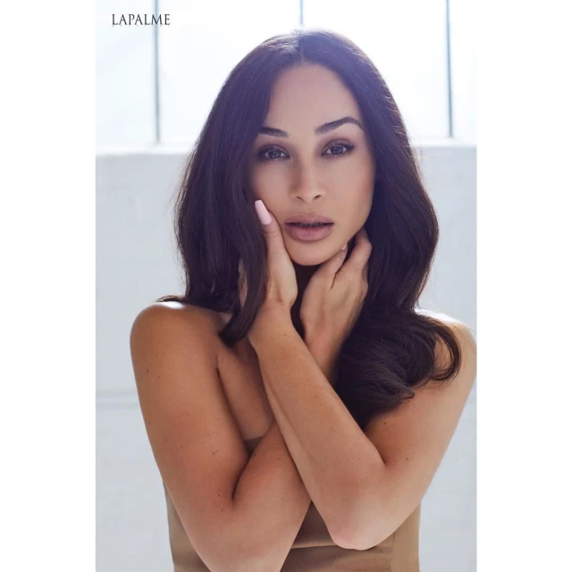Close-up studio shot of a brunette adult female with hands on her face, wearing a nude top against a bright neutral background.