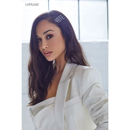 Portrait of a woman with dark hair wearing a white blazer and a hair clip that reads 'VOTE', posing against a neutral background.