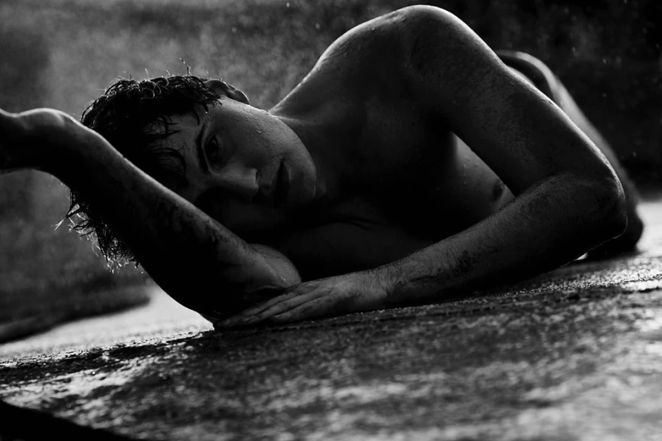 Monochrome portrait of a young adult male lying on a stone surface in a shallow depth of field with water droplets in the air.