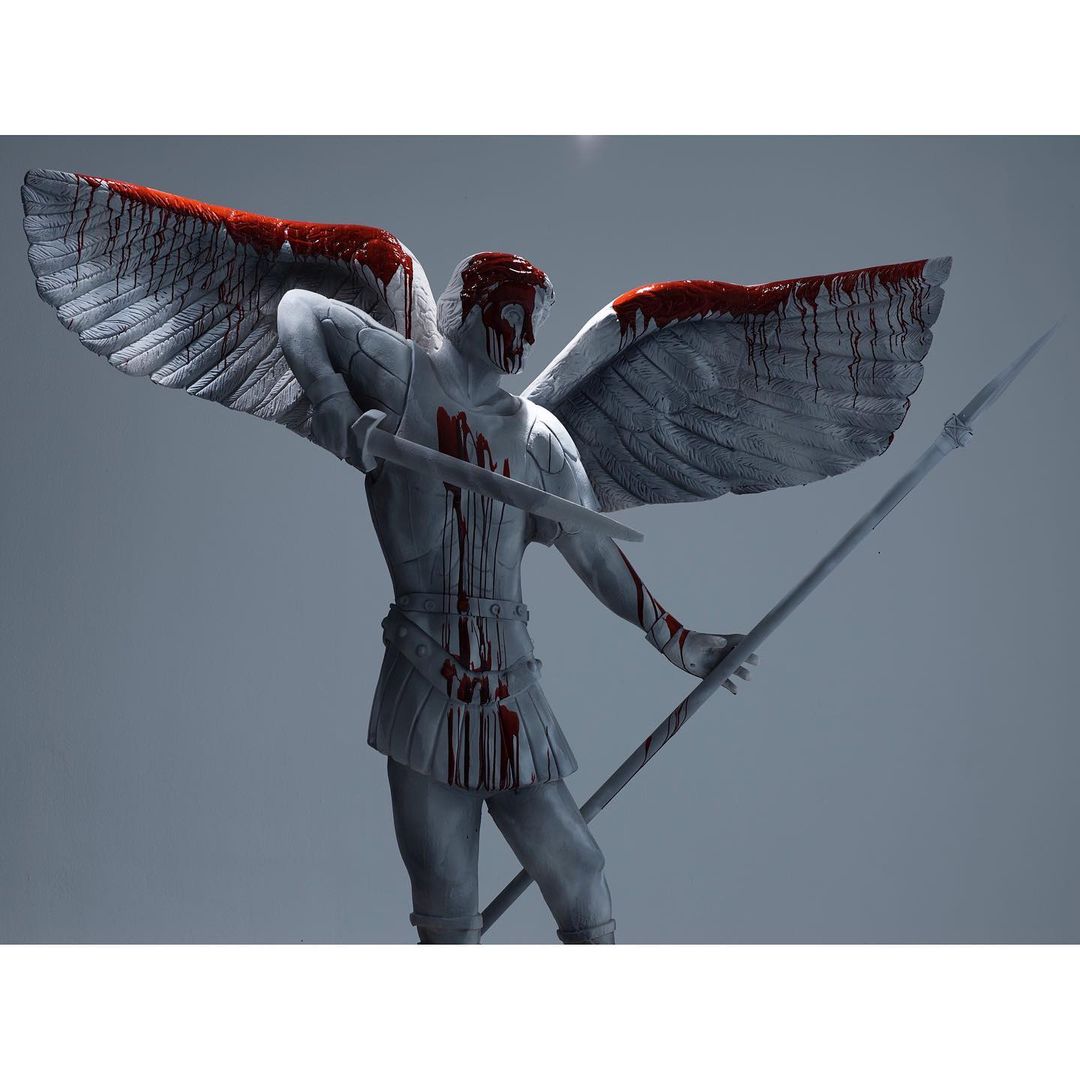 Grey-skinned angel statue with wings spread holds a spear and sword, covered in red paint on a solid grey background.