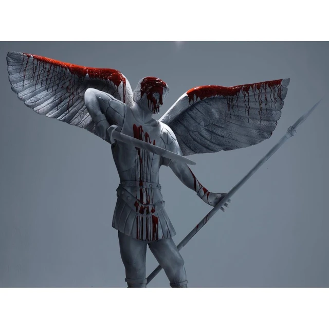 Grey-skinned angel statue with wings spread holds a spear and sword, covered in red paint on a solid grey background.
