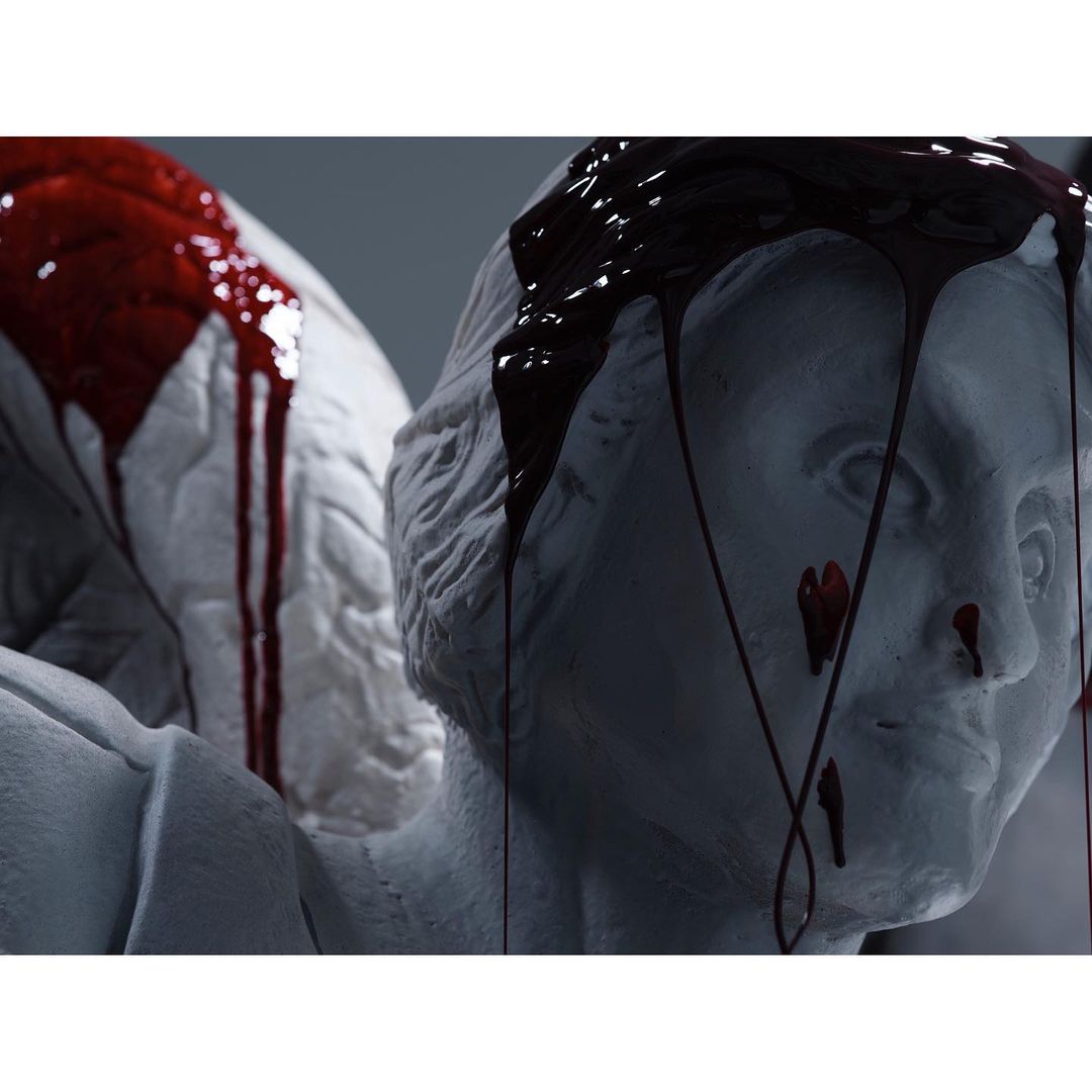 Close-up of two white classical sculpture heads with dark red liquid dripping down their faces in an indoor setting.