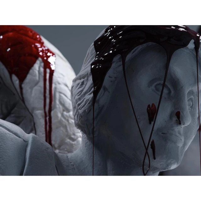 Close-up of two white classical sculpture heads with dark red liquid dripping down their faces in an indoor setting.