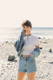 Smiling woman in white t-shirt and denim shorts holding denim jacket on a sandy beach