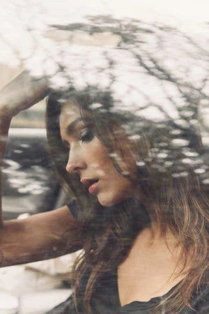 Portrait of a woman viewed through a textured glass pane reflecting tree branches, captured by photographer Rowan Daly.