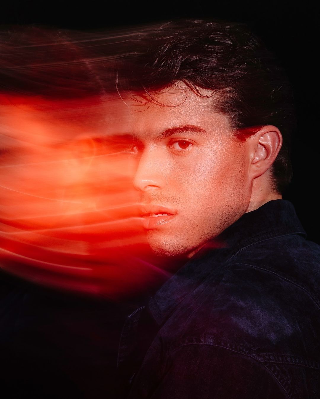 Portrait of a handsome adult male with dark hair, wearing a dark blue jacket, against a dark background with red light trails.