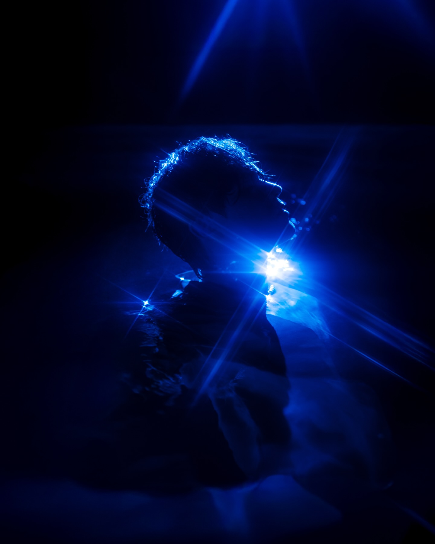 Silhouette of a person against a dark background, illuminated by a bright blue light creating lens flares and a dramatic, high-contrast image.