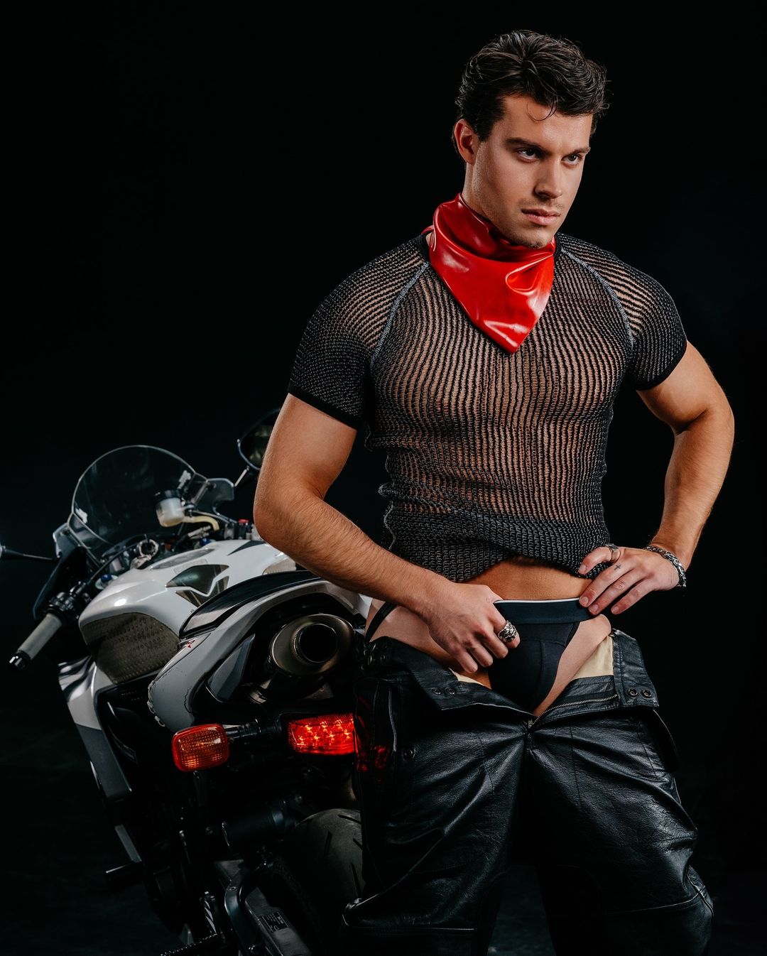 Masculine adult male poses next to a white motorcycle in a photo studio, wearing an open pair of leather pants and a red neckerchief.