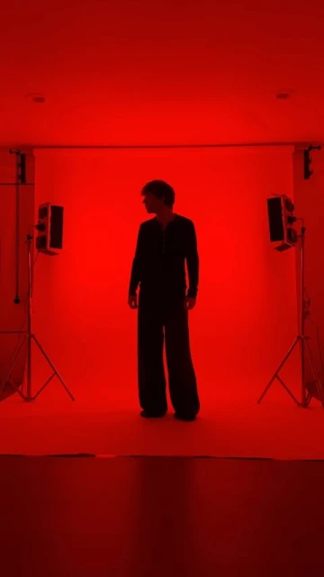 Full body shot of a person in wide leg suit standing in photo studio with red lighting and two light stands in the background.