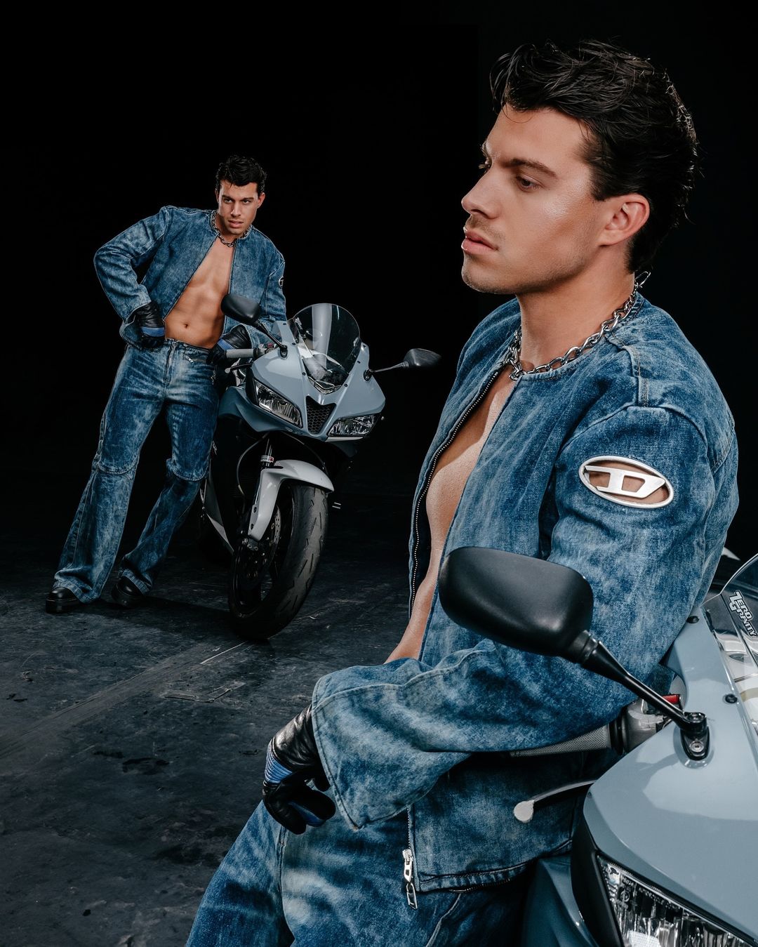 Two adult males pose in denim next to a grey motorcycle in a studio. One man looks off to the side; the other stands with hands on the bike.