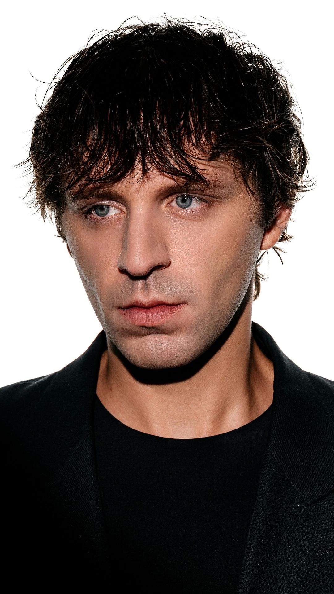 Headshot of a dark-haired adult male in a black jacket against a white background, looking off to the side with a serious expression.