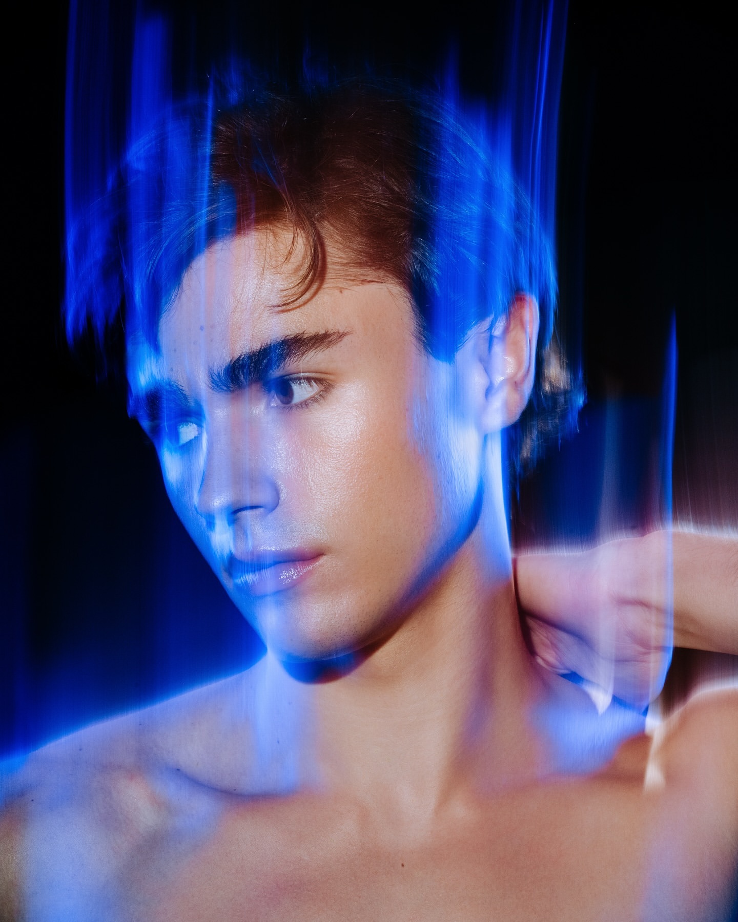 Portrait of a young adult male with a light complexion and dark hair, illuminated by blue light against a dark background in a studio.
