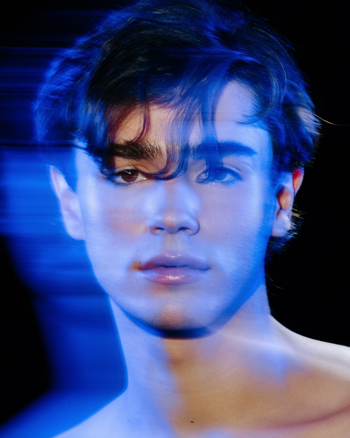 Close-up portrait of a young adult male with dark hair and light skin, accented with blue light and a black background.